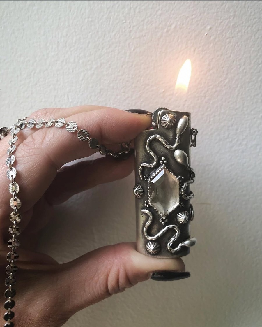 A hand holding a sterling serpent FireKeeper lighter case, lit, against a white wall