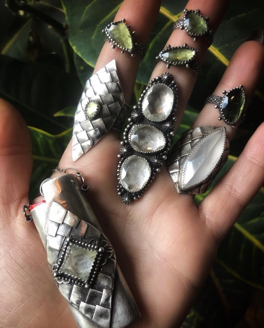 An open hand wearing a full set of large sterling rings with green and clear stones, holding a woven FireKeeper case, over green leaves