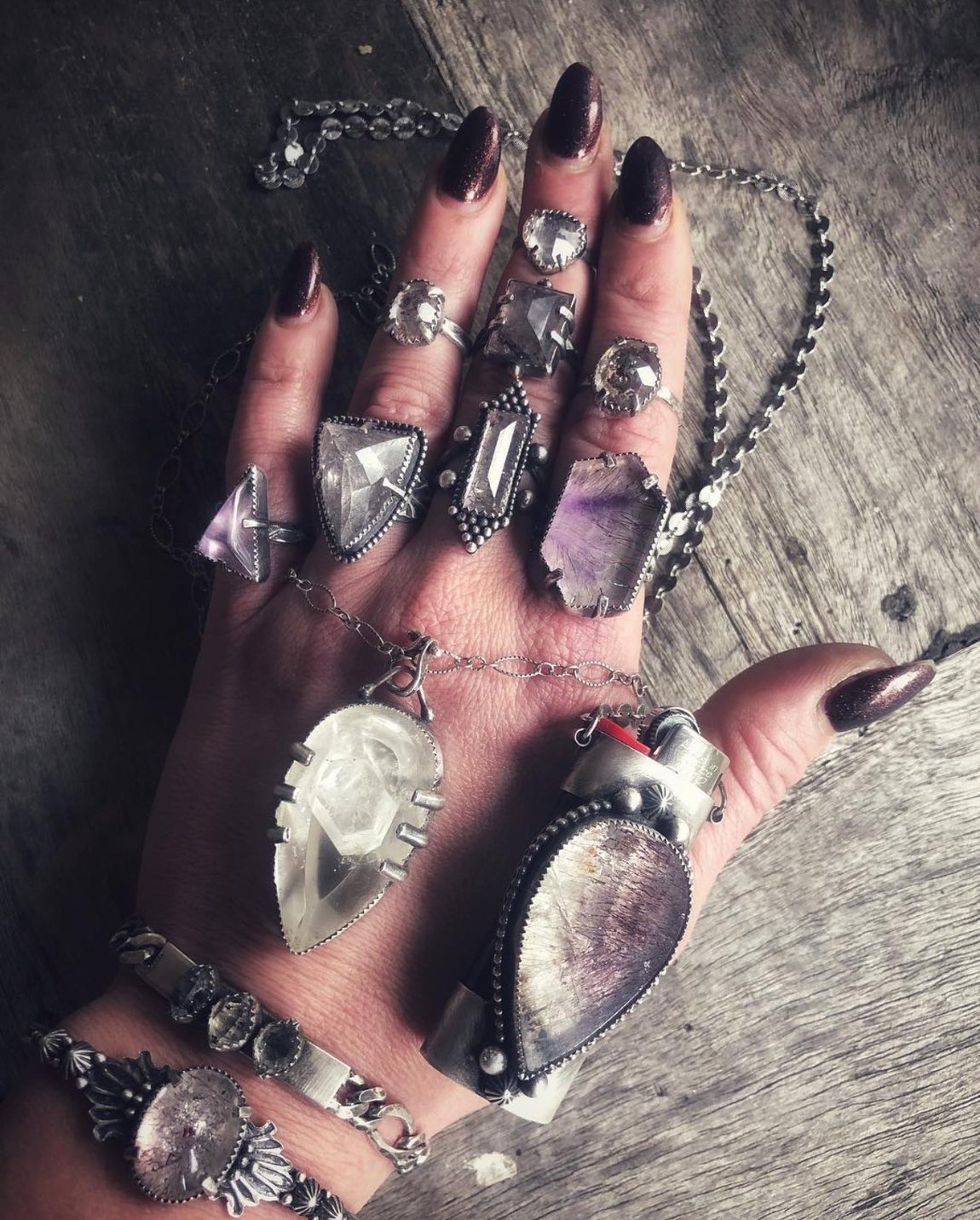 Jenna's open palm wearing a full set of her hand-forged rings, a FireKeeper lighter case and stacked stone bracelets, on weathered wood